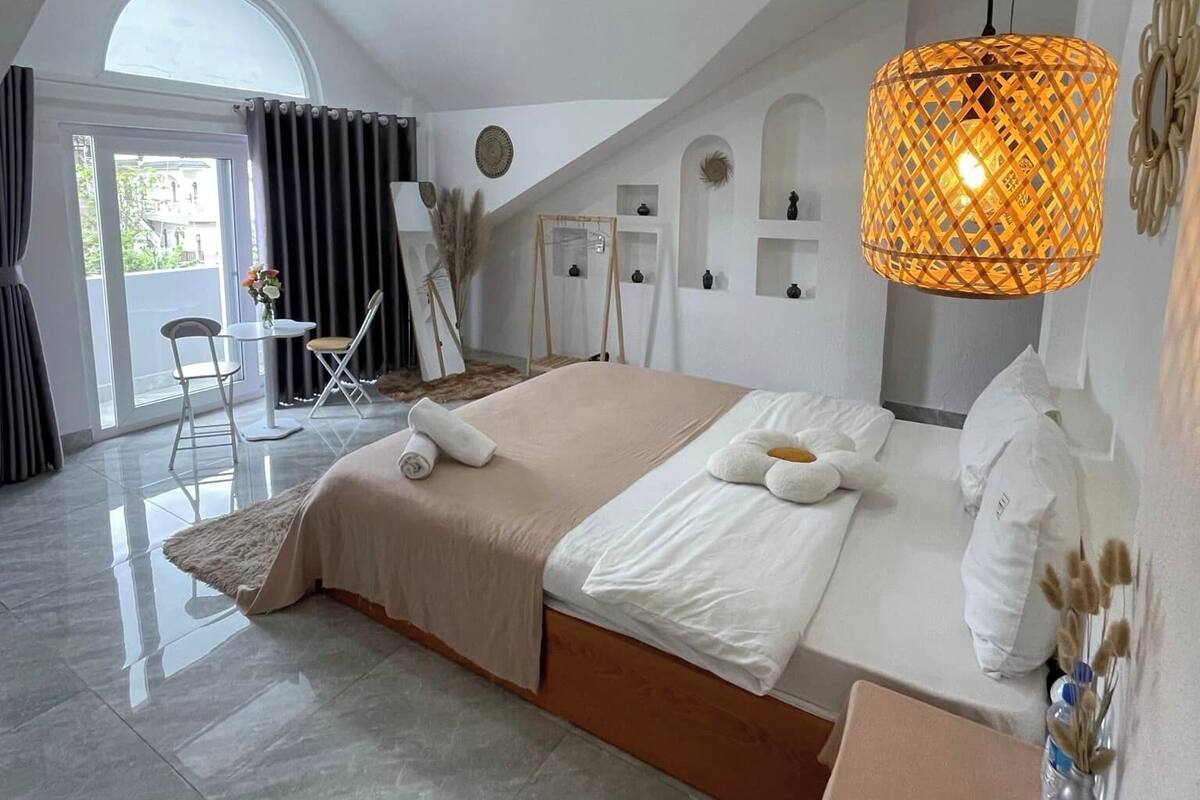Een moderne slaapkamer met een groot bed, een zithoek met een tafel en stoelen, een hanglamp en uitzicht door een groot raam.