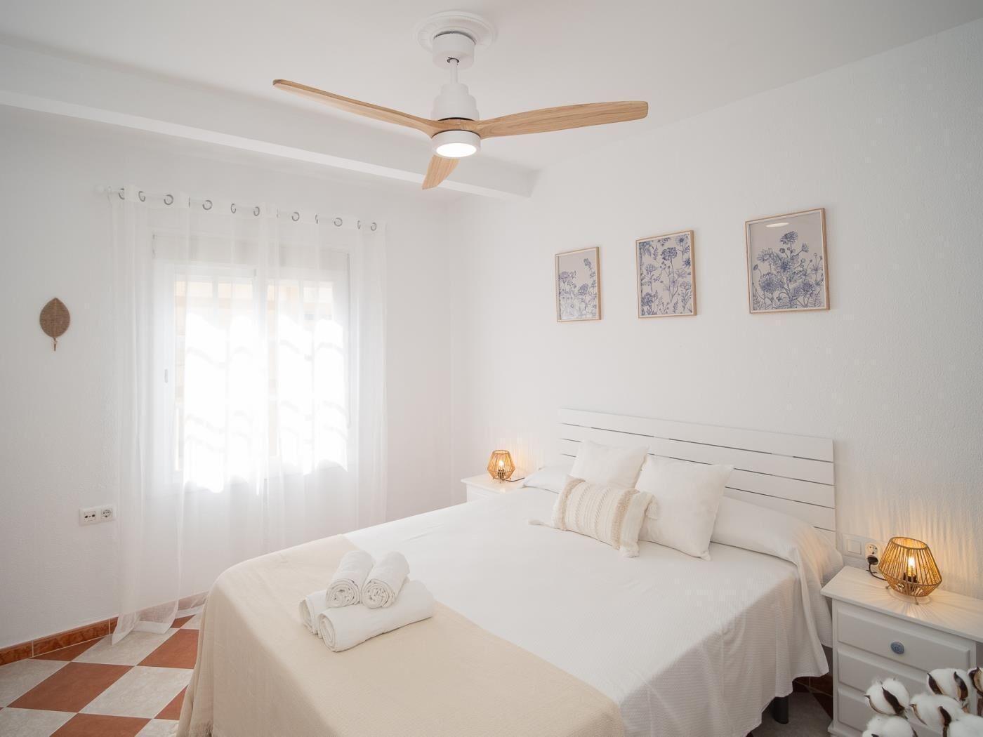 Une chambre d’hôtel avec un grand lit, un ventilateur de plafond, une table de chevet avec une lampe et trois tableaux représentant des motifs floraux, accrochés au mur.