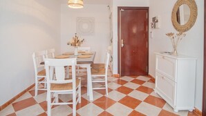 Een eetgedeelte met een geruit vloertje, witte stoelen, een tafel met placemats, een hanglamp, een houten deur en een witte console met een spiegel.