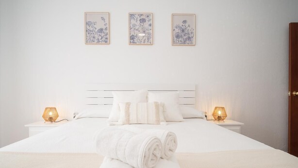 Een net opgemaakt bed met opgerolde handdoeken ervoor, een wit hoofdeinde en drie ingekaderde bloemwerken aan de muur.