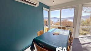 Ein Zimmer mit Tisch und Stühlen, großen Fenstern mit Blick auf ein Gewässer und Hügel sowie einer Klimaanlage an der Wand.