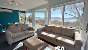 Ein Wohnzimmer mit zwei beigefarbenen Sofas, einem Glastisch und Blick auf einen See und Berge durch große Fenster.