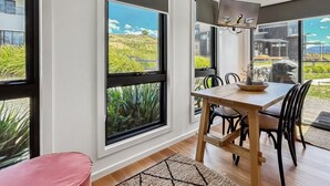 Comedor con mesa y sillas de madera, ventanas amplias que ofrecen vista a un paisaje con césped y edificios.