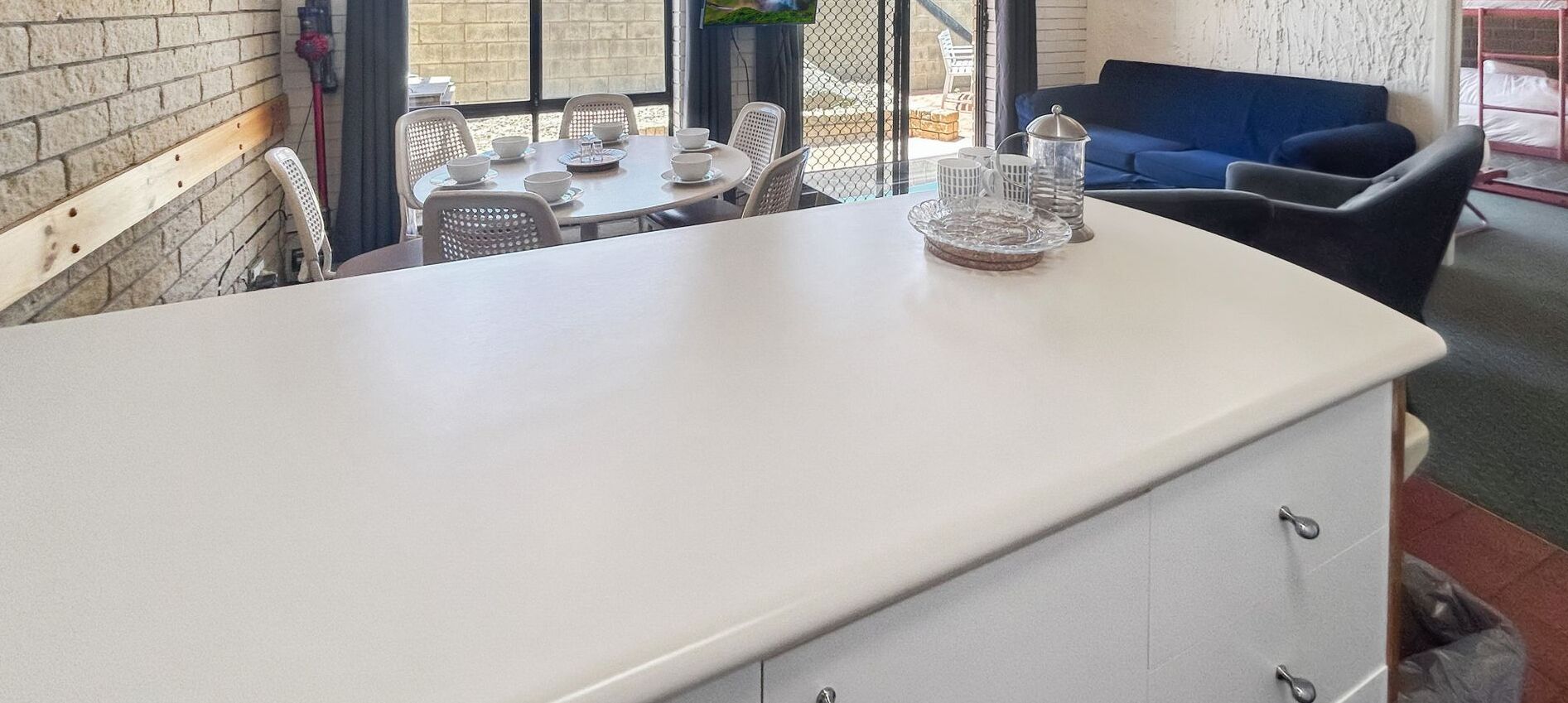 A kitchen area with a white countertop, a dining table set with white chairs, and a blue sofa in the background.