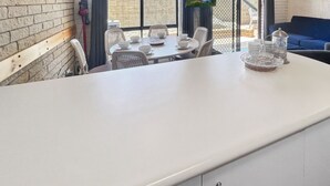 A kitchen area with a white countertop, a dining table set with white chairs, and a blue sofa in the background.