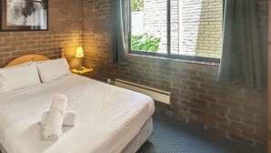A neatly made bed with white linens and a rolled towel, a wooden headboard, a nightstand with a lamp, and a window with blinds.