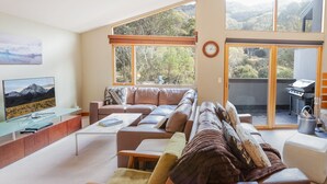 A spacious living room with a large window, a flat-screen TV, and a barbecue grill outside. - Escape 2 - Crackenback Ridge (Crackenback Ridge)