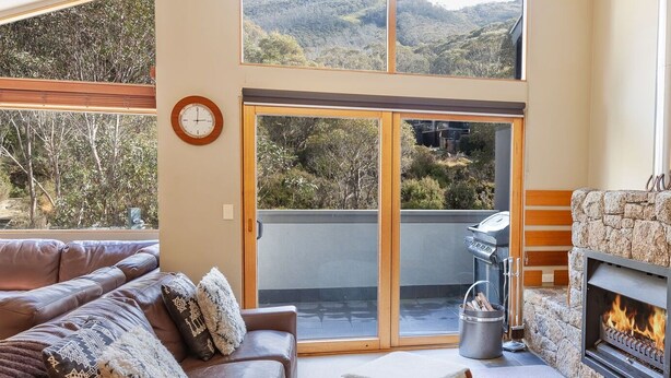 A cozy living room with a leather couch, a fireplace, and a large sliding glass door leading to a balcony with a scenic view. - Escape 2 - Crackenback Ridge (Crackenback Ridge)