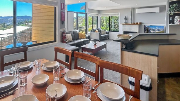A dining area with a view of the city, a living room with a sofa and chairs, and a kitchen with a TV. - Dromaius 7 amazing lake and mountain views with double garage (Jindabyne)