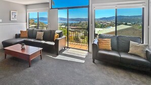 A living room with a sofa set, a coffee table, and a view of a coastal landscape through large windows. - Dromaius 7 amazing lake and mountain views with double garage (Jindabyne)