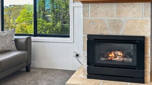 A fireplace with a stone surround, a leather sofa, and a window with a view of trees. - Dromaius 7 amazing lake and mountain views with double garage (Jindabyne)