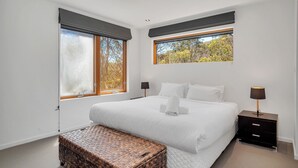 Une chambre avec un grand lit, deux tables de chevet, une lampe de chevet, un banc en osier et une fenêtre donnant sur des arbres.