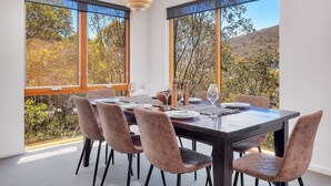 Un coin repas avec une table dressée pour six personnes, de grandes fenêtres et une vue sur les arbres et les collines.