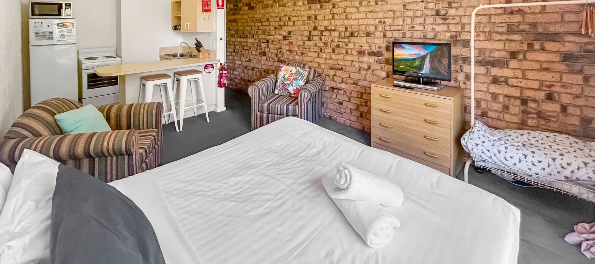 Une chambre d’hôtel avec un lit, une télévision, un bureau avec un micro-ondes, une petite kitchenette et un mur en briques.