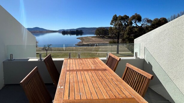A balcony with a wooden table and chairs overlooking a lake and distant hills. - Horizons 317 studio right on the Lake (Jindabyne)