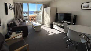 A modern hotel room with a sofa, a dining table, a TV, and a view of a lake and mountains. - Horizons 317 studio right on the Lake (Jindabyne)