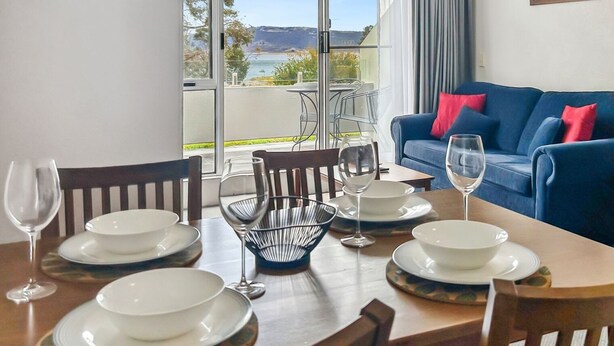 Un coin repas avec une table dressée pour quatre personnes, un canapé bleu agrémenté de coussins rouges et une vue sur l’extérieur grâce à de grandes fenêtres.