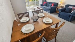 Un coin repas avec une table en bois dressée pour quatre personnes, entourée de chaises en bois. On aperçoit, à l’arrière-plan, un canapé bleu et une petite table d’appoint avec une lampe.