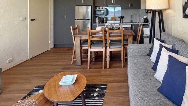 A modern living room with a grey sofa, wooden dining table, and chairs. A kitchen is visible in the background. - Alpine Mountain View 9 (Jindabyne)