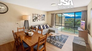 A modern living room with a dining area, a sofa set, a large window with a view, and artwork on the wall. - Alpine Mountain View 9 (Jindabyne)