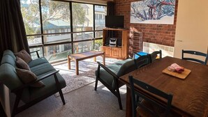 Un salon confortable avec un canapé, une petite table et une table à manger avec des chaises. On y trouve un téléviseur posé sur un meuble en bois, ainsi qu’un tableau accroché au mur.