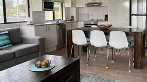 A compact living space with a sofa, a dining table with four chairs, a kitchen area with modern appliances, and a large window providing a view of the outdoors. - Alpine Mountain View 41 (Jindabyne)