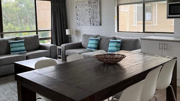 A modern living room with a dark wooden dining table, grey sofas with striped cushions, a white chair, and a kitchen area with a microwave and cabinets. - Alpine Mountain View 41 (Jindabyne)