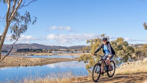 A cyclist on a dirt path beside a lake with hills in the background. - Alpine Mountain View 41 (Jindabyne)