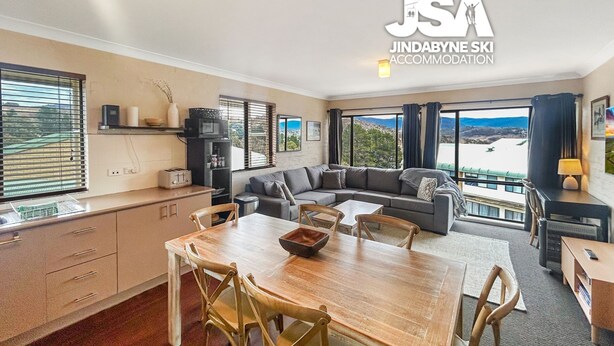 A modern living room with a wooden dining table, grey sofa, and large windows offering a view of the mountains.