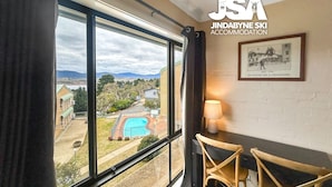 A room with a large window overlooking a swimming pool and a scenic view of hills and buildings. There is a framed picture on the wall and a lamp on a table.