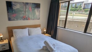 A hotel room with a bed, two bedside tables, a window with a view of buildings, and abstract art on the wall. - Alpine Mountain View 47 (Jindabyne)
