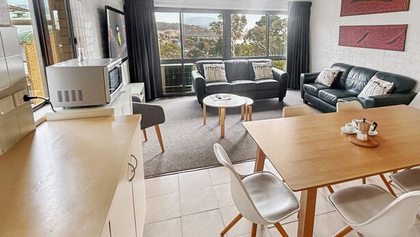 A modern living room with a sofa, a dining table with chairs, and a kitchen area with a microwave. - Alpine Mountain View 47 (Jindabyne)