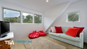 Ein Zimmer mit Fernseher, zwei Fenstern, einer Couch mit roten Kissen und einer roten Tasche auf dem Boden.