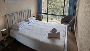 Ein ordentlich bezogenes Bett mit weißen Leinen und einem gerollten Handtuch, eine Nachttischlampe sowie Blick auf einen Swimmingpool durch ein großes Fenster.