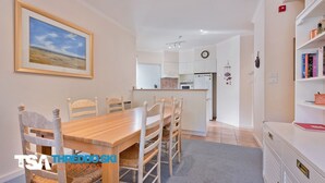 A dining area with a wooden table and chairs, a kitchen in the background, and a bookshelf with books. - Kaella - Central Village (Central Village)