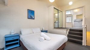 Hotellihuone, jossa on sänky, yöpöytä, sininen lipasto, kylpyhuone, jossa on pesuallas ja peili, sekä portaikko.