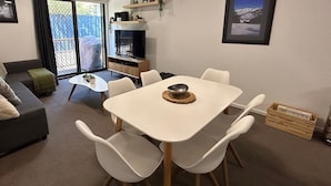 Un salon avec un canapé gris, une table à manger blanche avec quatre chaises, un téléviseur et une porte-fenêtre coulissante donnant sur un espace extérieur.