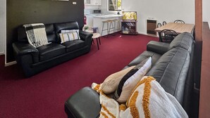 A living room with a black leather sofa, a red carpet, a dining area with a wooden table and chairs, a kitchen with white cabinets, and a sign that reads "PRAY FOR SNOW".