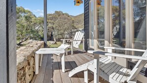 Eine Holzterrasse mit weißen Adirondack-Stühlen und einer Glastür, die zu einem Garten führt.