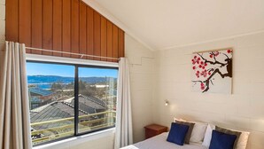 A bedroom with a large window, a bed with pillows, a small bedside table, and a wall-mounted artwork. - Moonbah 2 two bedroom unit (Jindabyne)