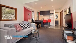 Ein modernes Wohnzimmer mit einer Couch, einem Couchtisch und einem Fernseher. Es gibt eine Küchenzeile mit einem Esstisch und Stühlen.
