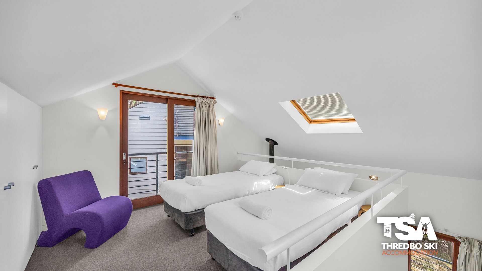A hotel room with a bed, a purple chair, a window with curtains, and a skylight.