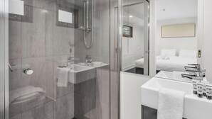 Ein modernes Badezimmer mit einer Duschkabine, einem Waschbecken mit Pflegeprodukten und einem Bett im angrenzenden Zimmer.