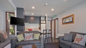 A modern living room with grey sofas, a flat-screen TV, and a kitchen area with a microwave and refrigerator.