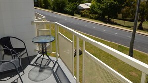 A balcony with a table and two chairs, overlooking a road with trees and a building in the background.