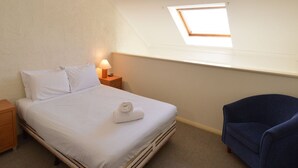 A single bed with white linens, a wooden nightstand with a lamp, a blue chair, and a skylight in a small room.