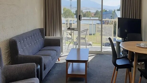 A living area with a sofa, a coffee table, and a TV. There is a balcony with a view of the outdoors.