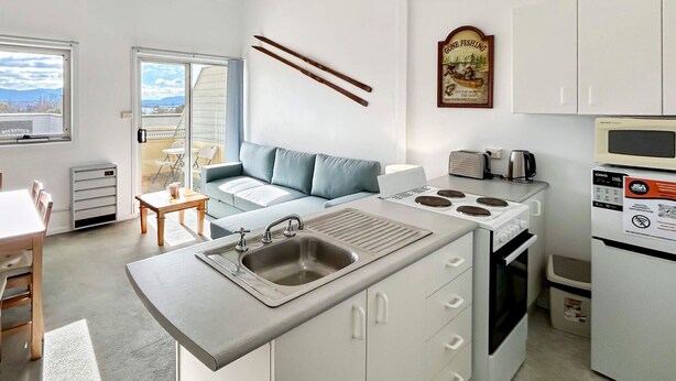 A compact kitchen with a built-in stove, a sink, and a microwave. There is a dining area with a table and chairs, and a sofa in the background. - Razorback 4 - Centrally located, self-contained 1 bedroom (Jindabyne)