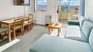 A modern living room with a wooden dining table and chairs, a light blue sofa, and a view of the outdoors through a large window. - Razorback 4 - Centrally located, self-contained 1 bedroom (Jindabyne)