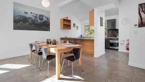 A modern dining area with a wooden table and chairs, a kitchen in the background, and a large painting on the wall.